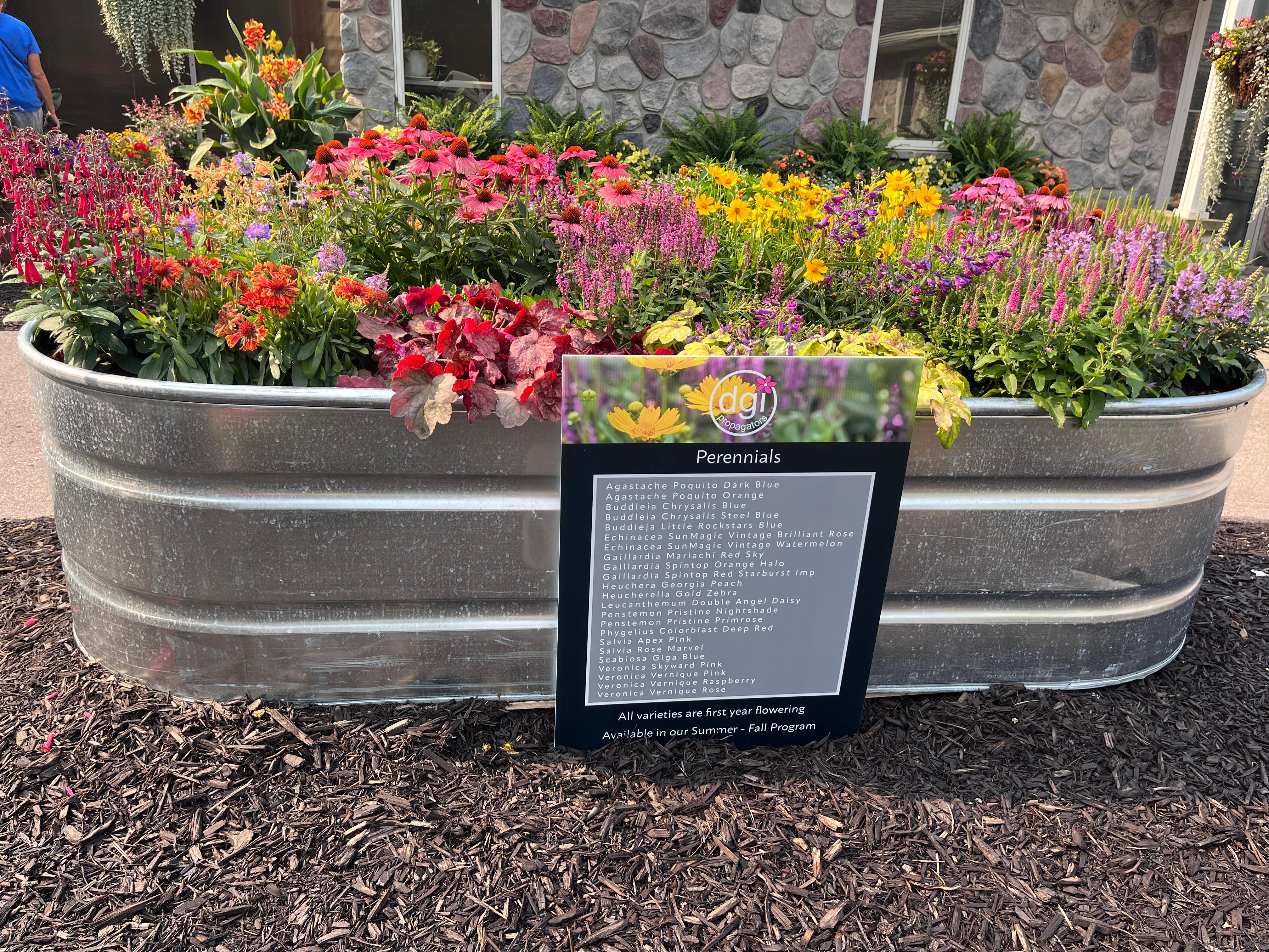 A large galvanized metal planter filled with a colorful mix of perennial flowers, including pink, purple, orange, yellow, and red blooms. In front, a sign lists the perennial varieties featured.